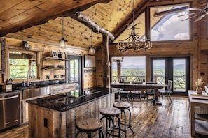 A rustic cabin interior with a large wooden dining area, a kitchen with a central island, and a view of the mountains through the windows. - Epic Mtn Views • Hot Tub • Fire Pit • Sleeps 13 (Blue Ridge)