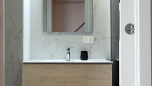 A bathroom with a marble wall, a white sink, a wooden vanity, and a mirror. - Hostmate - Bari - Garruba  (Bari)