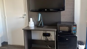 A room with a flat-screen TV mounted above a wooden desk, a small refrigerator, and a trash bin.