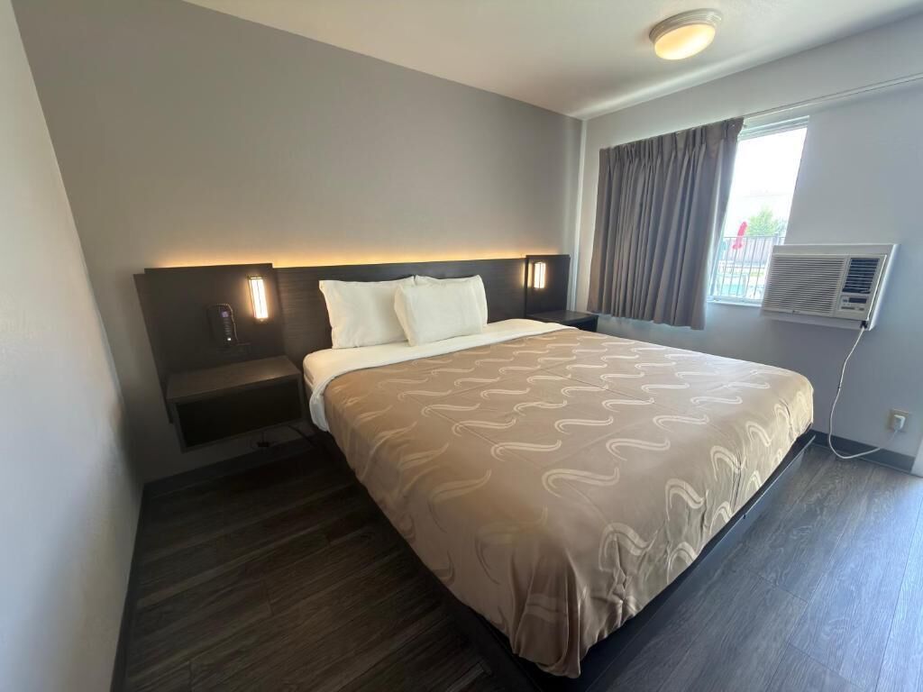 A hotel room with a bed, bedside tables, a wall-mounted air conditioner, and a window with curtains.