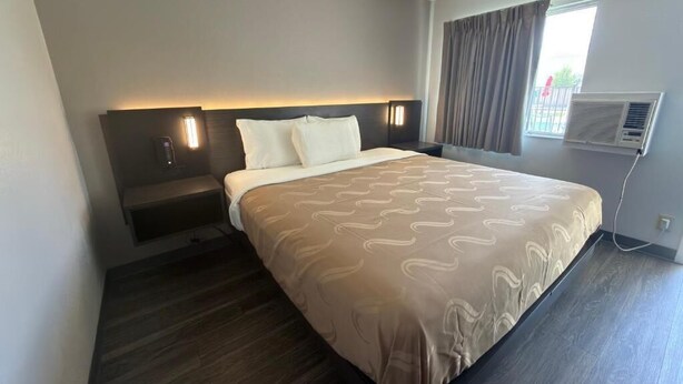 A hotel room with a bed, bedside tables, a wall-mounted air conditioner, and a window with curtains.