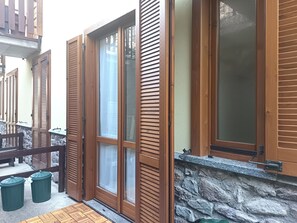Ein Fenster mit Holzzierleisten und Rollläden, eine Steinsäule und eine Holzterrasse mit Mülltonnen.