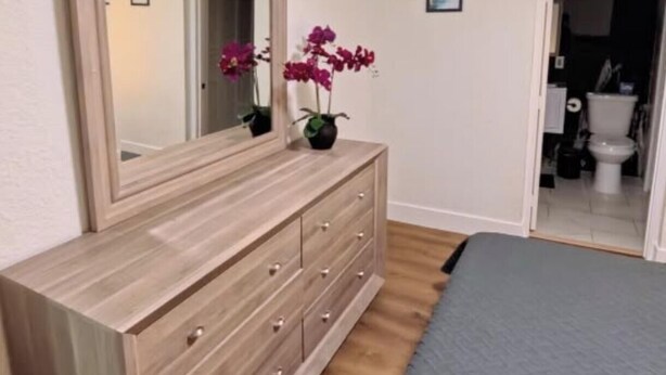 Een slaapkamer met een houten dressoir, een bed met grijze dekbedovertrek, een plafondventilator en een spiegel die een deur en een orchidee weerkaatst.