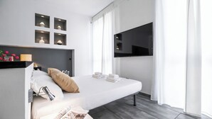 Ett modernt hotellrum med en säng, en platt-TV, ett nattduksbord med en lampa och ett fönster med gardiner.