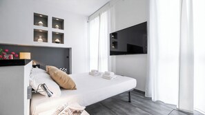 Ein modernes Hotelzimmer mit einem Bett, einem Flachbildfernseher, einem Nachttisch mit Lampe und einem Vorhangfenster.