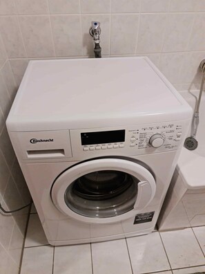 Una lavadora de carga frontal Bauknecht en un baño con suelo de baldosas y un cabezal de ducha.