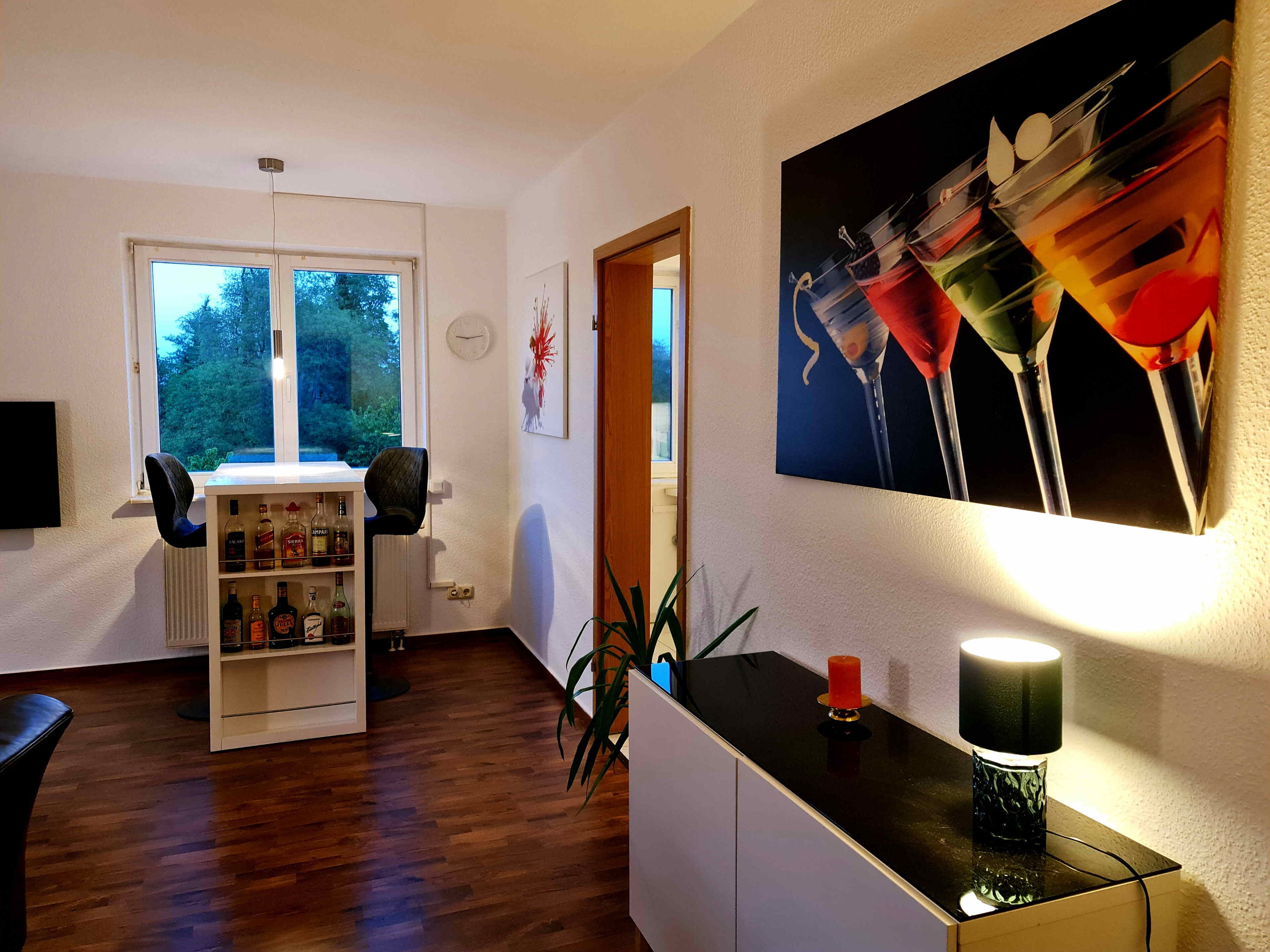 Een kamer met een houten vloer, een wit kastje, een bureau met een lamp, een boekenplank en een schilderij van glazen.