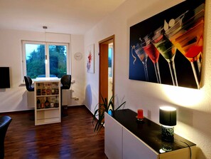 Una habitación con piso de madera, un armario blanco, un escritorio con lámpara, una estantería y un cuadro de vasos.