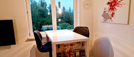 Un coin repas avec une table et des chaises, une fenêtre donnant sur un paysage verdoyant, une horloge murale et une étagère avec des bouteilles.