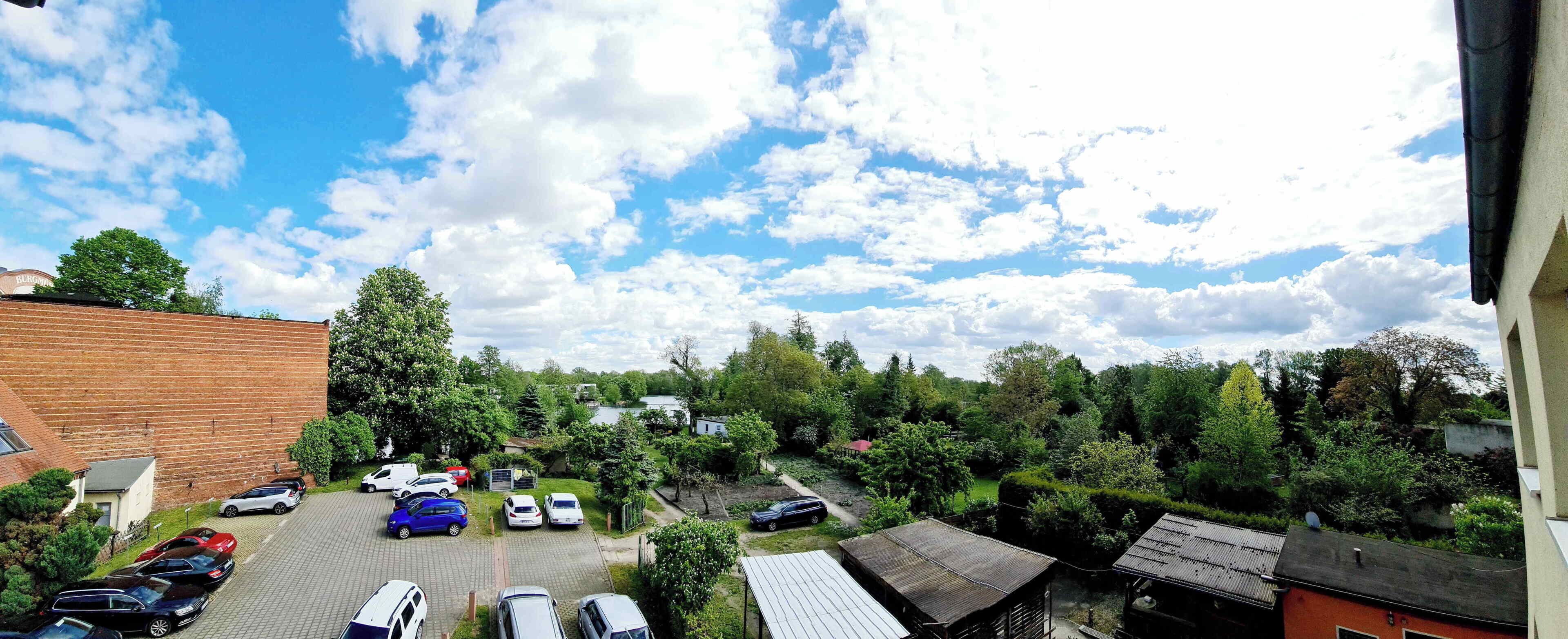 A parking area with multiple cars, a brick building, and a lush green landscape with trees and a body of water in the background.