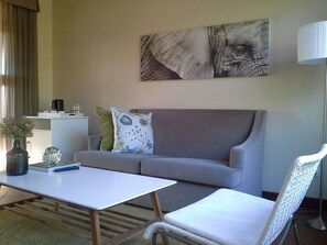 Una sala de estar con sofá, una mesita de centro, una silla, una lámpara, un cuadro y una pequeña mesa con un florero y un control remoto.