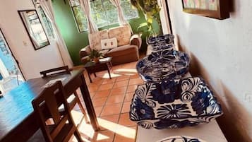 A dining area with a table, chairs, and a sofa. There is a window with curtains and a plant.