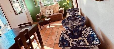 A dining area with a table, chairs, and a sofa. There is a window with curtains and a plant.