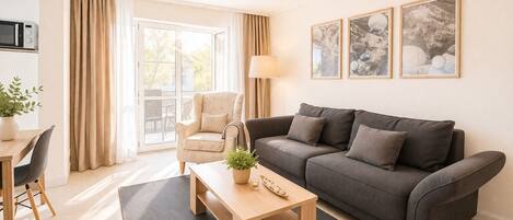 A modern living room with a sofa, a coffee table, a chair, and a lamp. There is a window with curtains, a plant, and framed pictures on the wall.