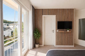 A bedroom with a large window, a potted plant, a TV mounted on a wooden panel wall, and a bed. - Villa Manatee - Wellness Apartment with a Balcony (Sellin)