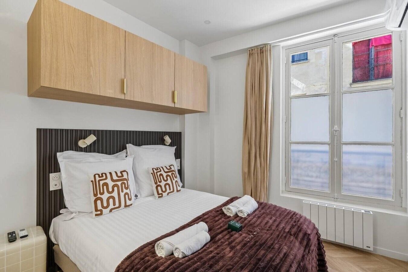 A hotel room with a bed, a window with curtains, a radiator, and a wooden wardrobe.