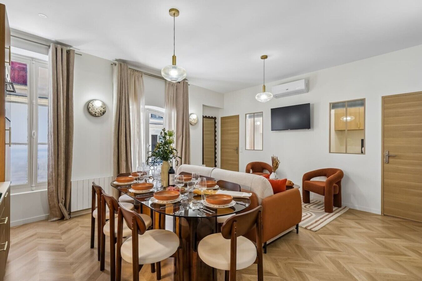 A dining area with a round table set for six, surrounded by wooden chairs. There is a sofa and a television mounted on the wall.