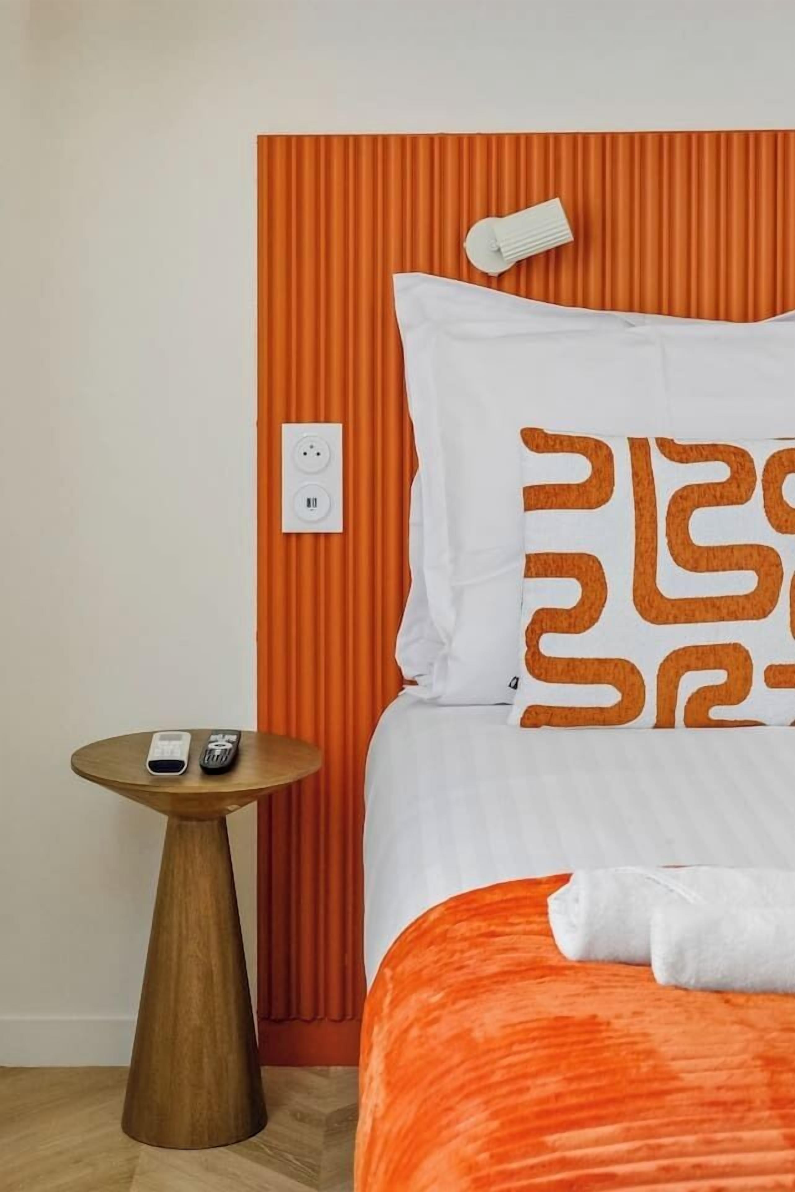 A hotel room with a bed, an orange headboard, a bedside table with a remote control, and a power outlet on the wall.