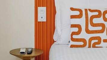 A hotel room with a bed, an orange headboard, a bedside table with a remote control, and a power outlet on the wall.