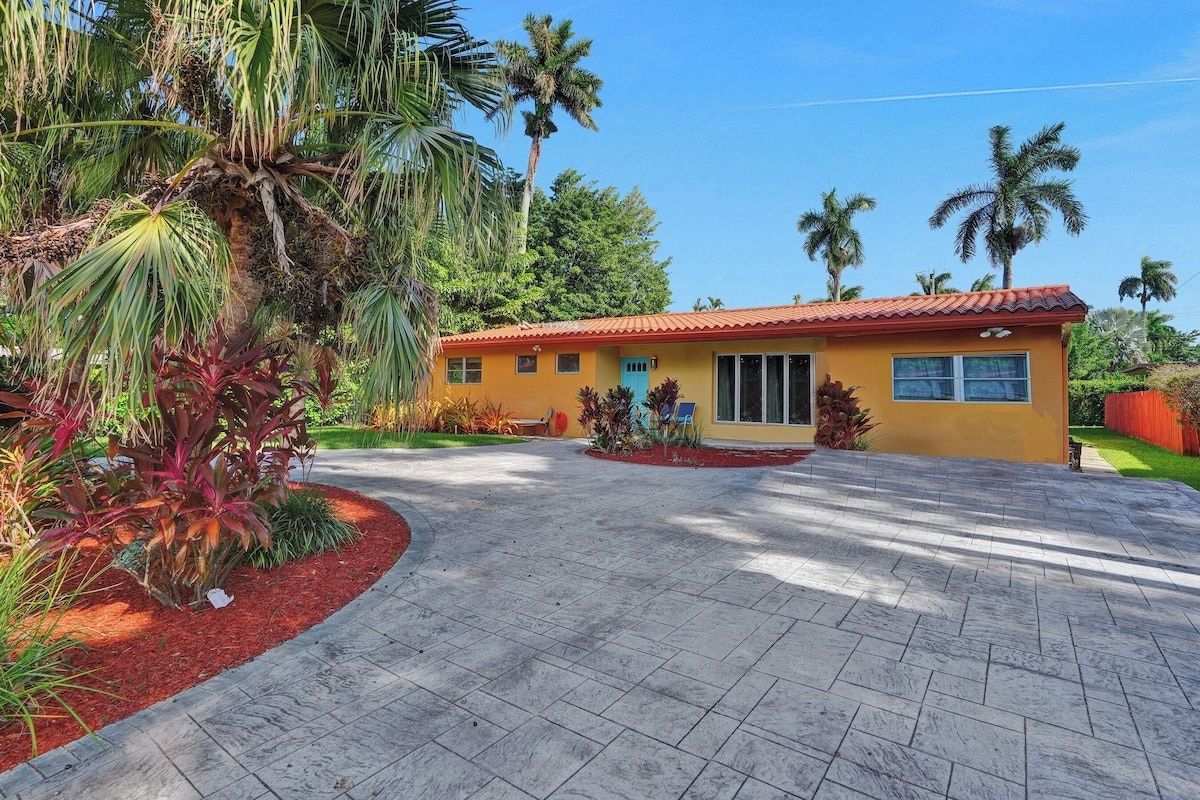 A yellow building with a red roof, surrounded by a landscaped garden with palm trees and red mulch.