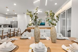 A modern dining area with a wooden table set for a meal, two large vases with greenery, and a kitchen in the background. - 5 Bd / 4.5 Ba / Sleeps 14 / Windsor Cay Resort (Clermont)