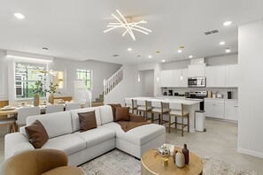 Uma sala de estar moderna com sofá seccional, área de jantar e cozinha com armários brancos e eletrodomésticos de aço inoxidável.