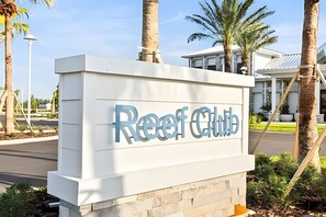 Une enseigne pour le Reef Club, au design moderne, située dans un espace paysager agrémenté de palmiers, avec un bâtiment en arrière-plan.