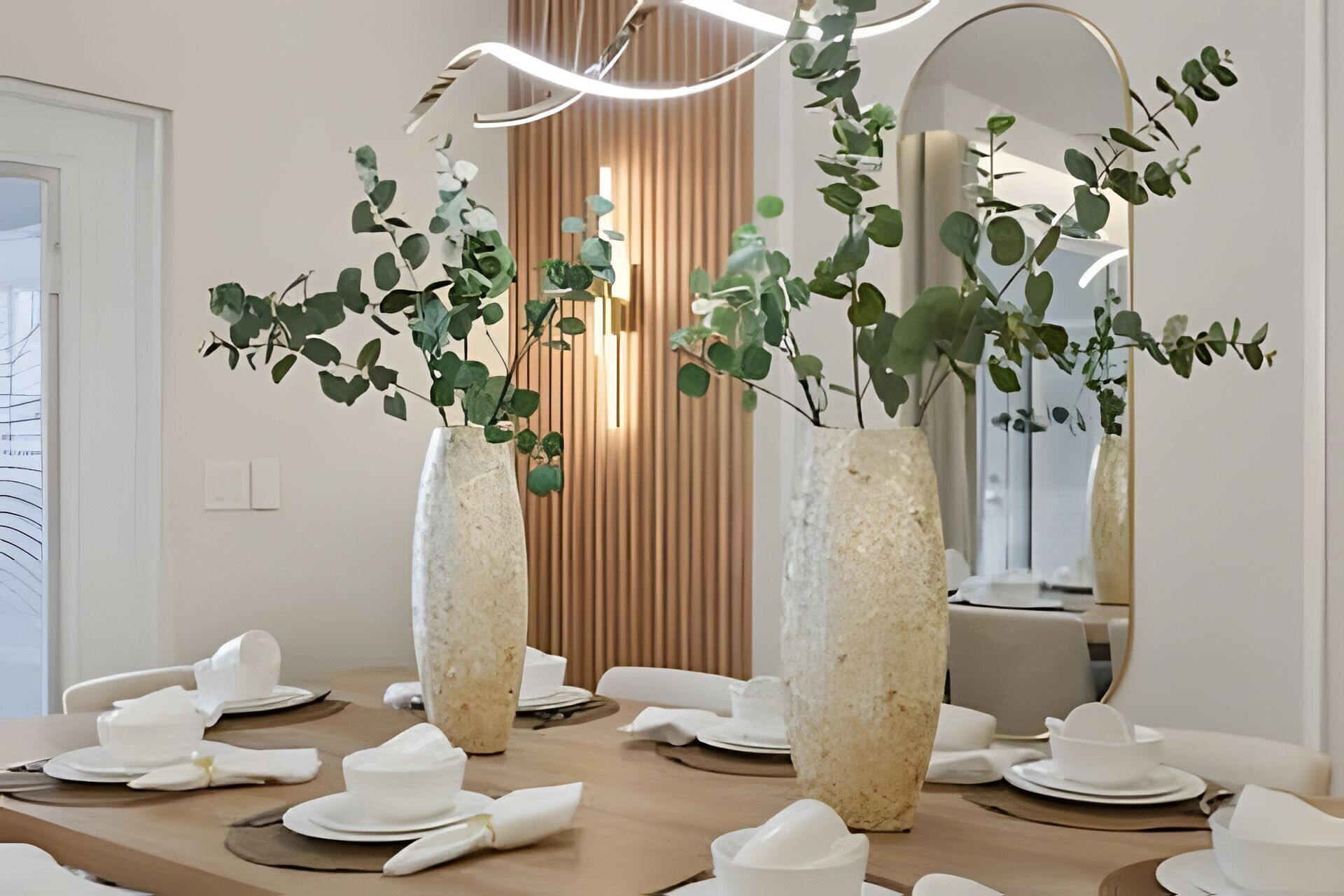 A dining table set with white tableware, flanked by tall, textured vases with green foliage.