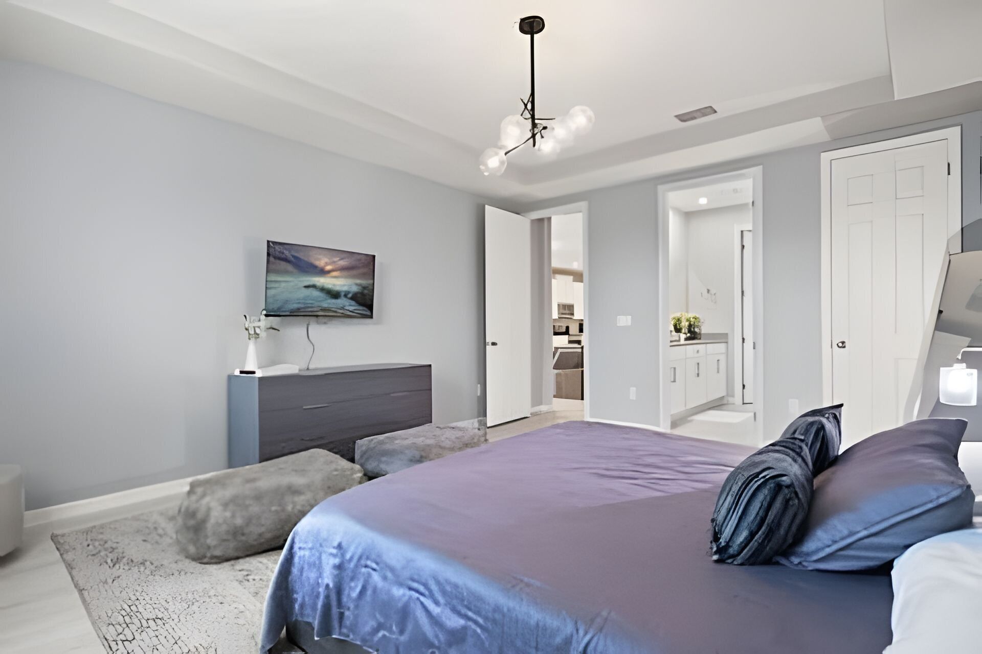 A modern bedroom with a bed, a dresser, a television, and a view into the kitchen.