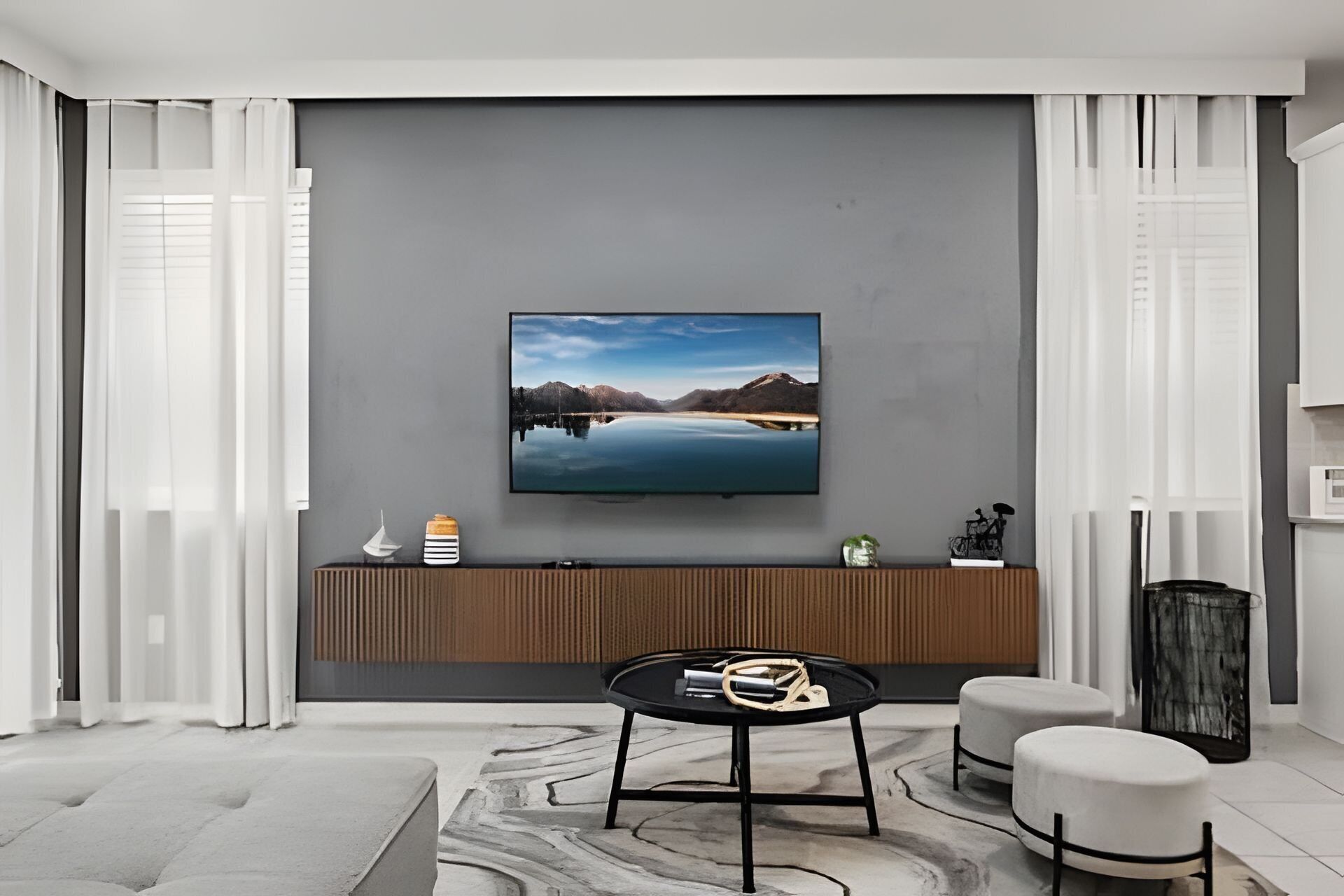 A modern living room with a flat-screen TV mounted on a grey wall, a wooden console table with decorative items, a round black coffee table, and two white cushioned stools.