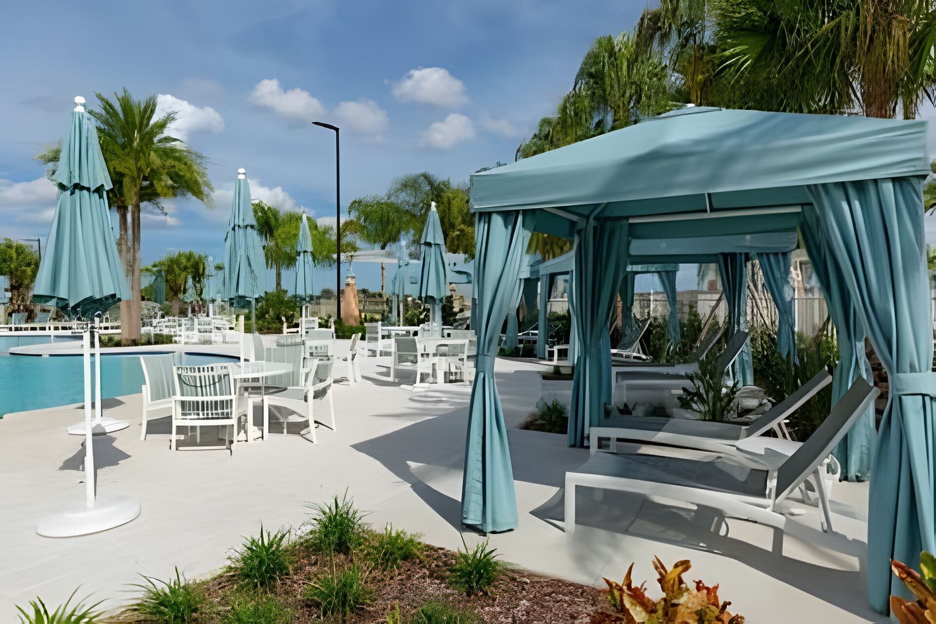 A poolside area with lounge chairs and umbrellas, surrounded by palm trees and a clear sky.