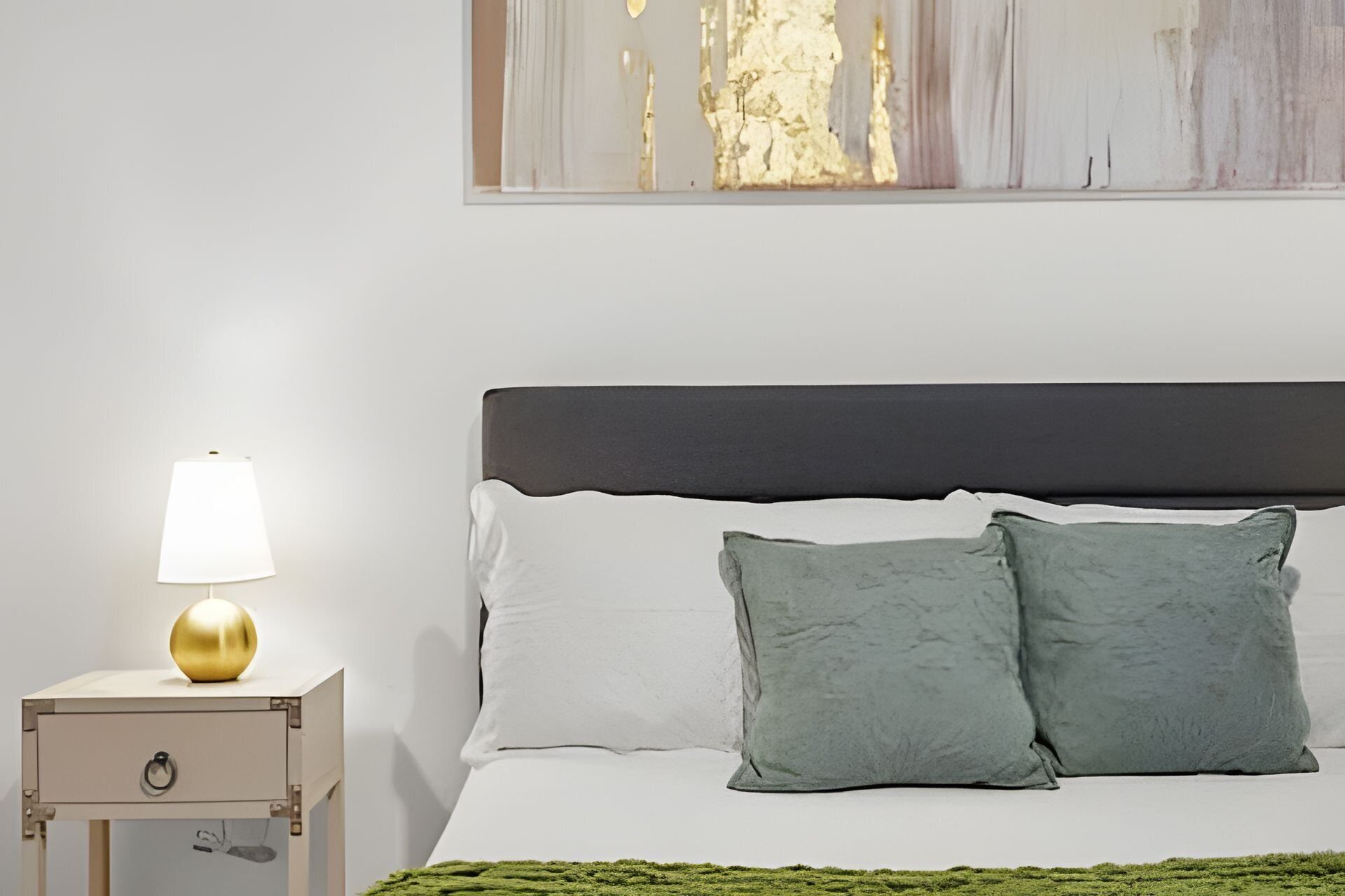 Una cama bien hecha con un cabecero oscuro, dos almohadas y una manta blanca. Se ve una mesita de noche con una lámpara y un cajón pequeño.