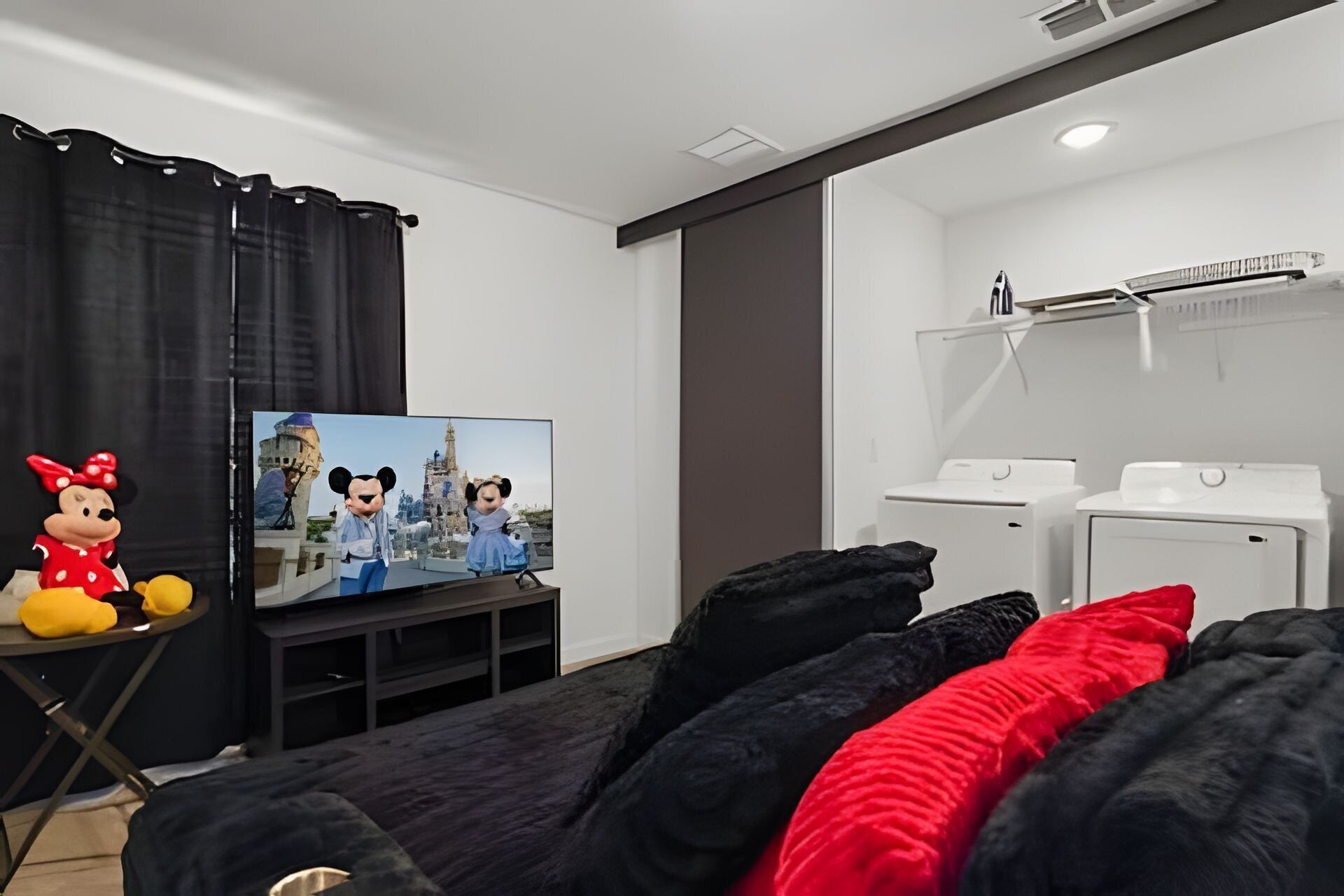 Quarto de hotel com televisão de tela plana, um boneco de pelúcia do Mickey Mouse, sofá preto e uma mesa pequena.