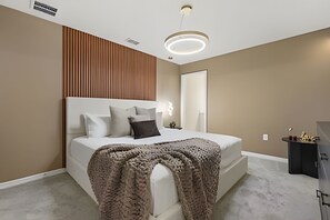 A modern bedroom with a large bed, a wooden panel wall, a round ceiling light, and a nightstand with a lamp.
