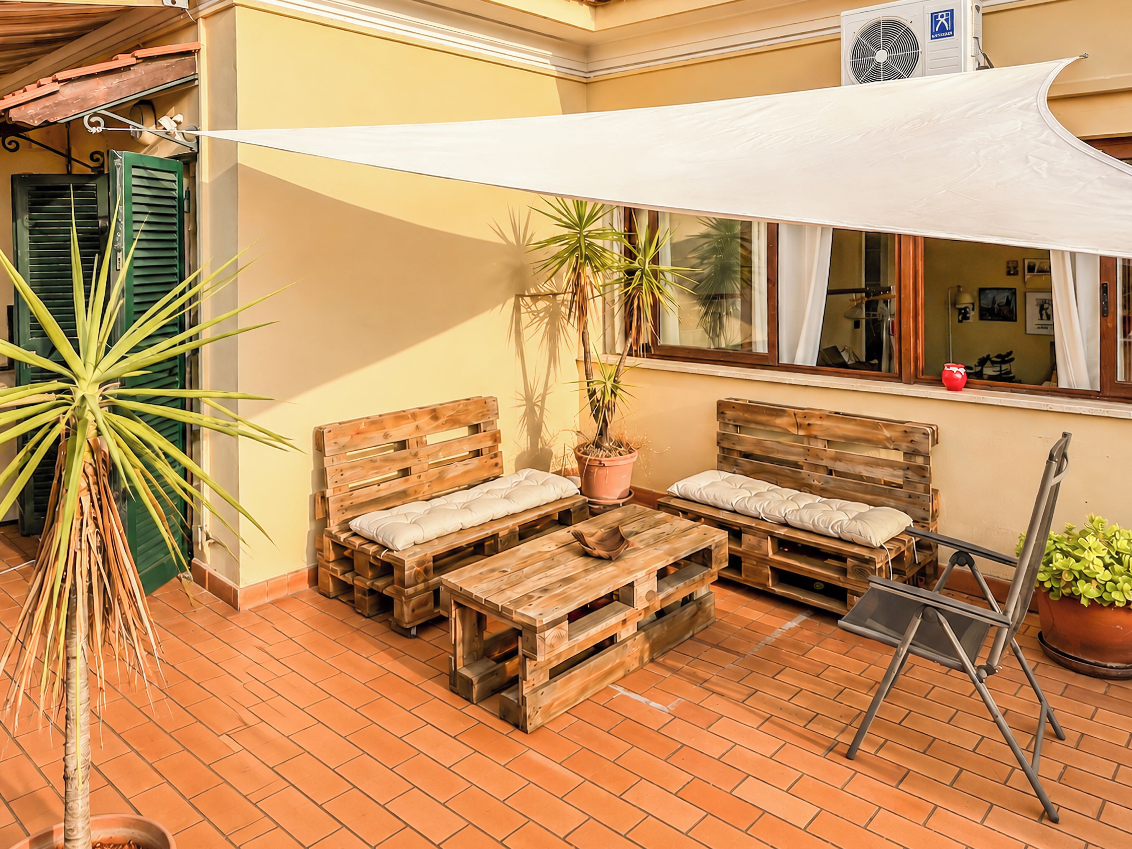 Un patio con mobili in legno, un tenda vela bianca, piante in vaso e pavimento piastrellato.