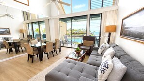 Uma sala ampla com um sofá cinzento grande, uma mesa de jantar com cadeiras, uma mesa de centro e vista para o oceano através de grandes janelas.