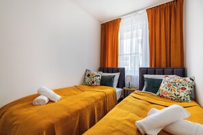 A hotel room with two beds, a nightstand, a lamp, and a window with curtains. - Czluchowska Apartament (Warszawa)