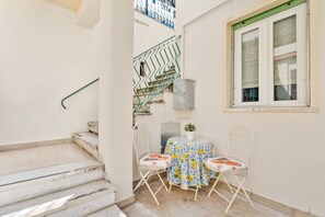 Ein kleiner Sitzbereich im Freien mit Tisch und zwei Stühlen, eine Treppe und ein Fenster mit Vorhang.