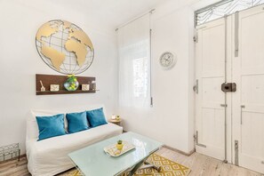 Une pièce avec un canapé blanc, une table basse en verre, une carte du monde accrochée au mur, une horloge et une porte blanche.