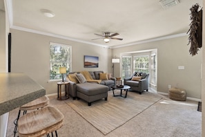 A living room with a grey sofa set, a coffee table, and a ceiling fan. There are two windows with blinds, a painting on the wall, and a small table with a lamp.