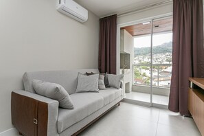 Ein modernes Wohnzimmer mit grauem Sofa, einem Holzschrank und Blick auf die Stadt durch eine Schiebetür.