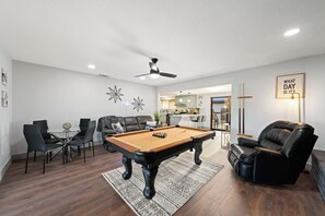 A modern living room with a pool table, leather chairs, and a kitchen in the background.