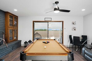 A pool table with a view of the outdoors, a ceiling fan, and a wooden cabinet with glass doors.