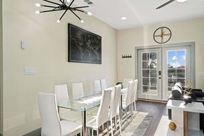 A dining area with a glass table and white chairs, a world map on the wall, a clock, and a door leading outside.