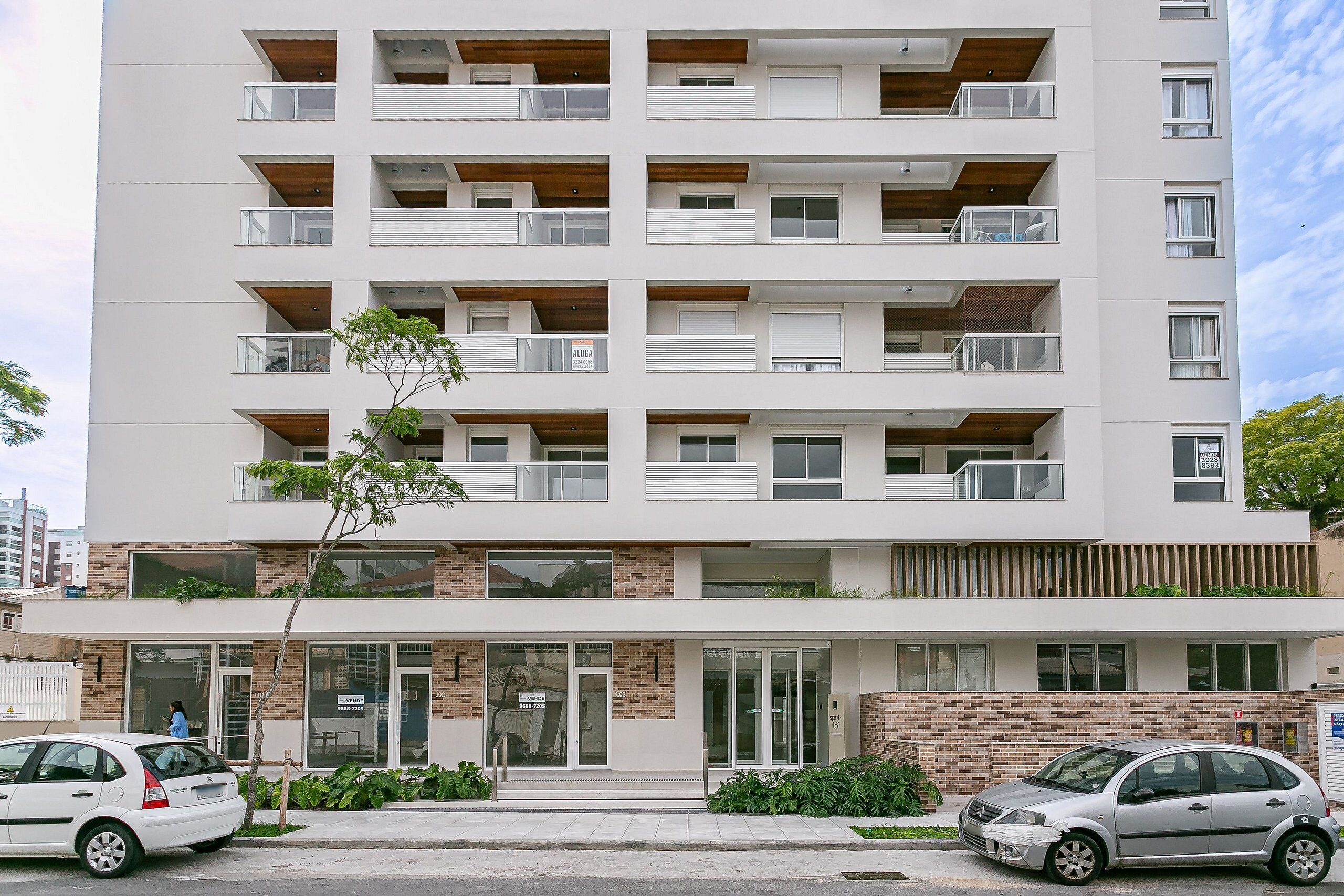 Edifício residencial de vários andares com varandas, entrada no nível da rua e placa 'VENDES' indicando imóvel à venda.