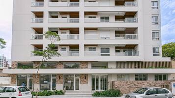 Edifício residencial de vários andares com varandas, entrada no nível da rua e placa 'VENDES' indicando imóvel à venda.