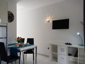 Ein modernes Zimmer mit Esstisch, Stühlen, einer Uhr, einem Fernseher und einem Schreibtisch mit Lampe.