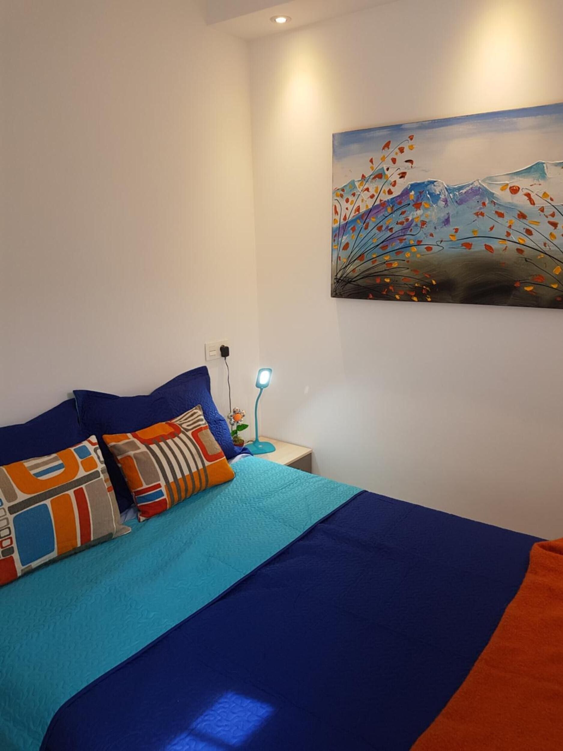 A bedroom with a bed, pillows, a nightstand, a lamp, and a painting on the wall.