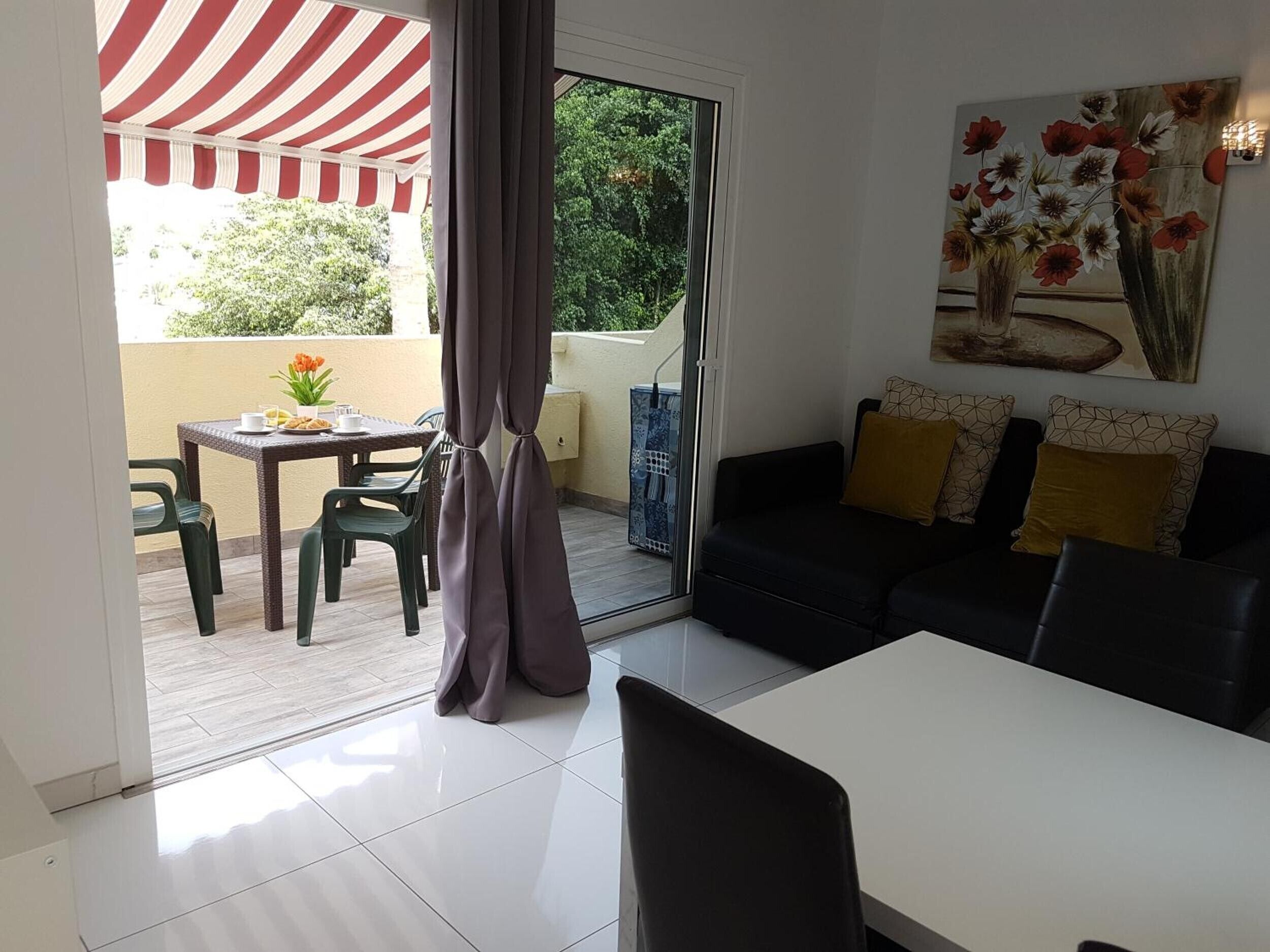A living room with a black sofa, a dining table, and a patio area with a striped awning.