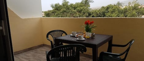 A balcony with a table set for two, chairs, a small plant, and a view of greenery and buildings.